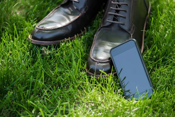 Vista de cerca del teléfono inteligente de arena de zapatos masculinos negros con pantalla en blanco en el césped verde - foto de stock