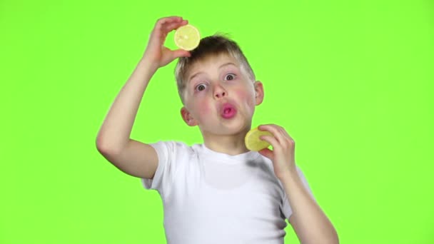 Мальчик с ломтиками лимона облизывает их и показывает гримасы. Зеленый экран. Медленное движение — стоковое видео