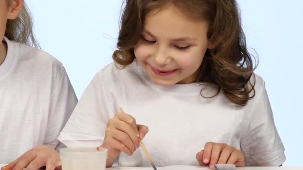 Los niños se sientan a la mesa y decoran los dibujos con colores. Fondo blanco. Movimiento lento — Vídeos de Stock