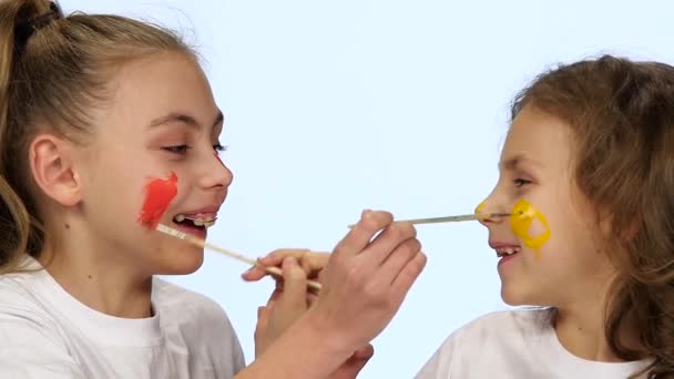 Kinderen zitten aan de tafel en zich op hun gezichten met verf penselen te schilderen. Witte achtergrond. Slow motion — Stockvideo