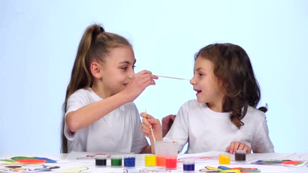 As crianças sentam-se à mesa e pintam-se em seus rostos com pincéis de tinta. Fundo branco. Movimento lento — Vídeo de Stock