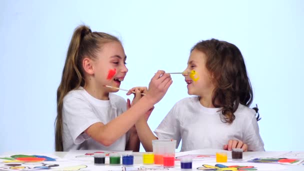 Dos niñas se sientan en la mesa, pintan las borlas en sus caras. Fondo blanco. Movimiento lento — Vídeos de Stock