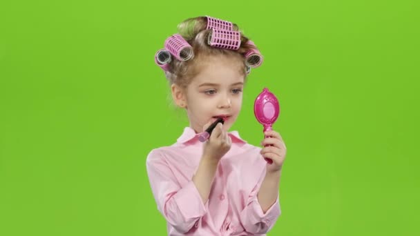 Kind in krulspelden houdt een spiegel in haar handen en schildert lippenstift met lippenstift. Groen scherm — Stockvideo
