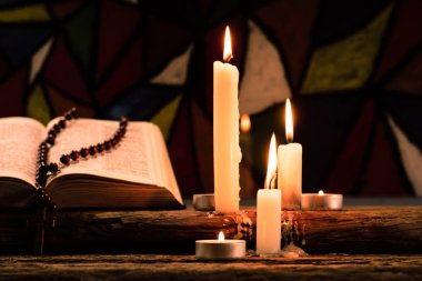 Bible crucifix and beads with a candle on a old oak wooden table.  Beautiful Stained-glass windows background.Religion concept. clipart