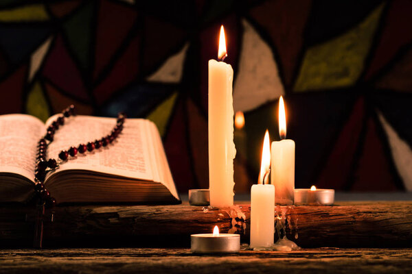 Bible crucifix and beads with a candle on a old oak wooden table.  Beautiful Stained-glass windows background.Religion concept.