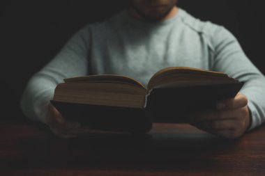 Genç adam kırmızı ahşap bir masada oturuyor. Eski kitabı ve siyah arka planı okuyor..