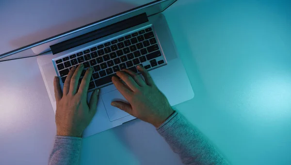 Man working night online silver laptop in the color light on a table night background. Top view.