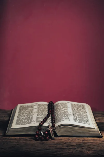 古いオークの木のテーブルの上で聖書とビーズの十字架を開きます 美しい赤い壁の背景 — ストック写真