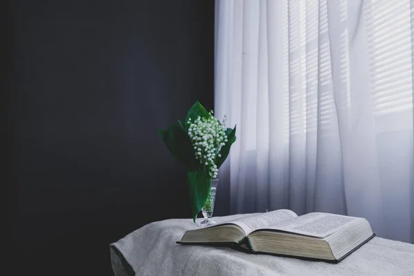 Open Holy Bible and flower in vase on a white table, black   wall and window  background. Stay at home. Religion concept.
