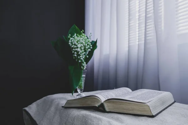 Open Holy Bible and flower in vase on a white table, black   wall and window  background. Stay at home. Religion concept.