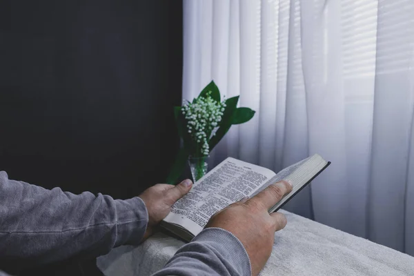 白いテーブル 黒い壁と窓の背景に花瓶に聖書と花を読んでいる男 家にいて 宗教概念 — ストック写真