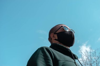 Yüz maskesi takan genç bir adam. Güneş gözlüklü yakışıklı adam siyah tıbbi maske takıyor, gökyüzü arkaplanı, fotokopi alanı. Salgın koronavirüs covid-19 karantina dönemi konsepti