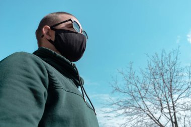 Yüz maskesi takan genç bir adam. Güneş gözlüklü yakışıklı adam siyah tıbbi maske takıyor, gökyüzü arkaplanı, fotokopi alanı. Salgın koronavirüs covid-19 karantina dönemi konsepti