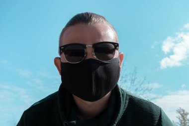 Yüz maskesi takan genç bir adam. Güneş gözlüklü yakışıklı adam siyah tıbbi maske takıyor, gökyüzü arkaplanı, fotokopi alanı. Salgın koronavirüs covid-19 karantina dönemi konsepti