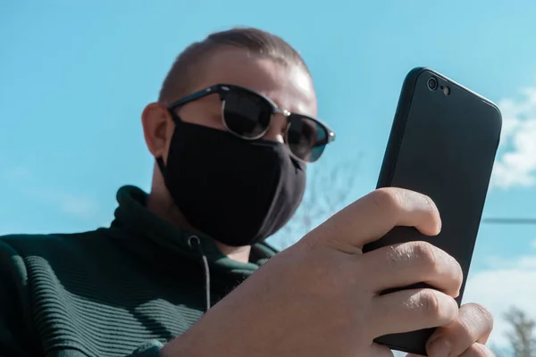 Young man wearing face mask. Handsome man in sunglasses wear black medical mask holding smartphone, sky background, copy space. Pandemic coronavirus covid-19 quarantine period concept.