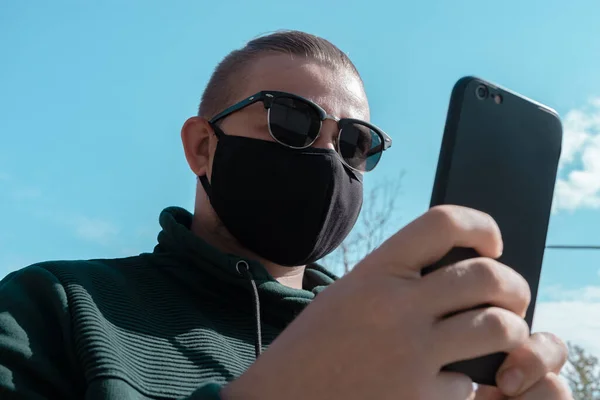 戴口罩的年轻人 戴着太阳镜的英俊男子戴着黑色医疗面罩 手持智能手机 天空背景 复制空间 大肠埃希菌Covid 19检疫期概念 — 图库照片