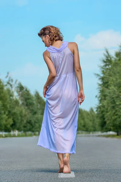 Güzel ince model kadın güneş, mavi gökyüzü ve yeşil ağaçları ile yolda arkadan hafifçe poz bir mor ipek elbise şeffaf kısa Kahverengi kıvırcık saçlı. Moda fotoğraf açık çekim