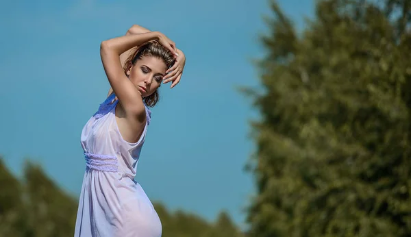 Güzel ince model kadın kısa kıvırcık kumral bir mor ipek elbise yavaşça elle parlak güneş, mavi gökyüzü ve yeşil ağaçlar ile açık poz. Moda portre kadın modeli fotoğraf çekim