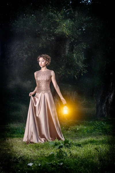 Lambaları altın uzun Abiye kısa Kahverengi kıvırcık saçlı güzel seksi ince kız ışık pozlar açık fairytale koyu yeşil ormanda. Kadın modeli ile fantezi sanat fotoğraf çekimleri