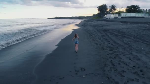 美丽快乐的年轻女性在牛仔裤短裤和灰色 T恤沿着海滩的海洋与黑色的沙子和波浪在巴厘岛日落 拍摄视频4K 从上面的飞机 美妙的景色 惊人的模型构成 — 图库视频影像