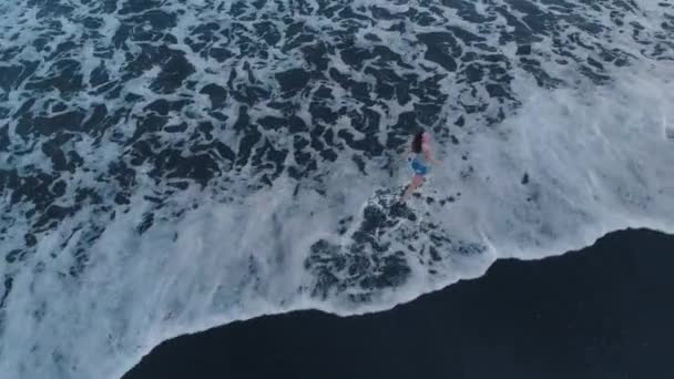 ジーンズのショート パンツ グレーの シャツは素晴らしいビュー上から航空機から のビデオを撮影し バリ島の日没時の黒い砂と波と海のビーチに沿って歩いて美しい幸せな若い女性 — ストック動画