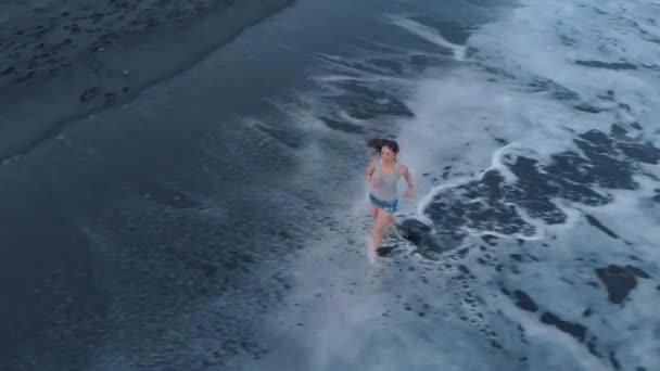 美丽快乐的年轻女性在牛仔裤短裤和灰色 T恤沿着海滩的海洋与黑色的沙子和波浪在巴厘岛日落 拍摄视频4K 从上面的飞机 美妙的景色 — 图库视频影像