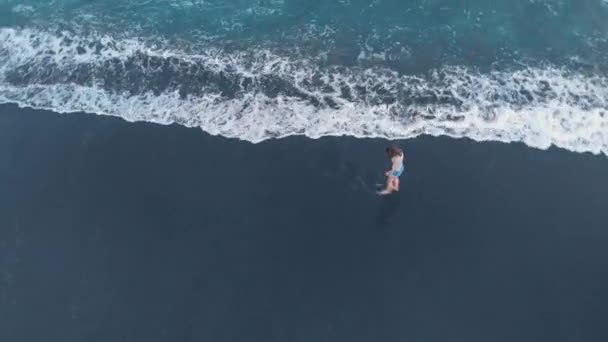 美丽快乐的年轻女性在牛仔裤短裤和灰色 T恤沿着海洋海滩漫步与黑色沙滩和波浪在巴厘岛日落 拍摄视频4K 从上面的飞机 美妙的景色 — 图库视频影像