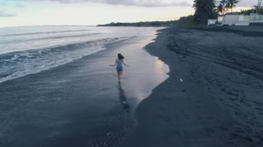 Siyah kum ve kot şortlu kadın modeli ve gri tişört koşma, video 4 k uçak yukarıdan, harika manzara çekim güzel mutlu genç ile Bali günbatımı, dalgalar okyanus plaj havadan görüntüleri