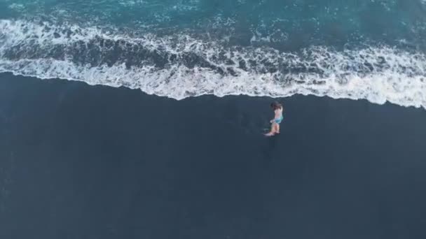 海洋海滩与黑色的沙子和波浪在巴厘岛的空中视频与美丽的快乐年轻的步行女性模特牛仔裤短裤和灰色 拍摄视频4K 从上面的飞机 美妙的景色 — 图库视频影像