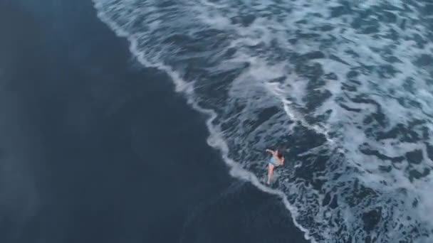 Images aériennes de la plage océanique avec du sable noir et des vagues au coucher du soleil sur Bali avec une belle jeune femme heureuse en short en jeans et un T-shirt gris posant sur la plage, tournage de la vidéo 4K de l'avion d'en haut, vue magnifique 
