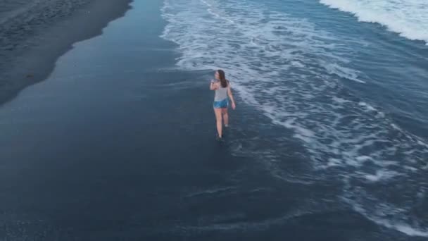 海洋海滩的空中视频与黑色的沙子和波浪在巴厘岛日落与美丽的快乐年轻的步行 运行女性模特牛仔裤短裤和灰色 拍摄视频4K 从上面的飞机 美妙的景色 空气基 — 图库视频影像