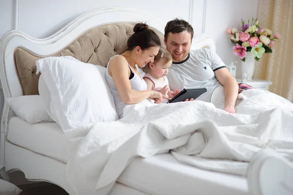 Young parents and their little daughter are looking at the tablet while lying in bed Stock Photo