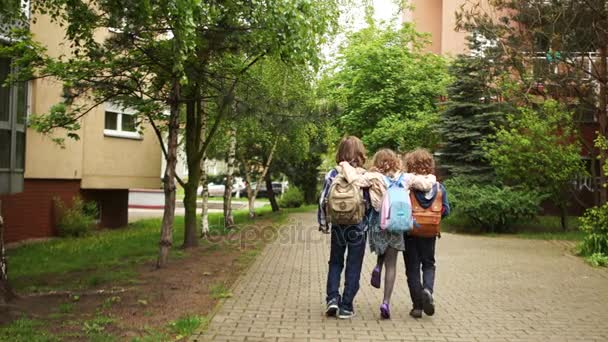 Tres escolares - una niña y dos niños vienen de la escuela abrazando. Secretos infantiles. Amistad escolar, primer amor . — Vídeos de Stock