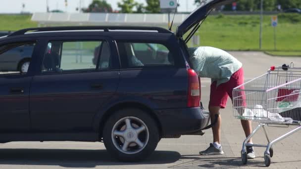 Un hombre joven y atractivo carga productos en el maletero de un coche desde un carro de supermercado. El papel de los hombres en la familia. Distribución de funciones. Canasta de comida. El domingo de compras. Problemas domésticos — Vídeo de stock