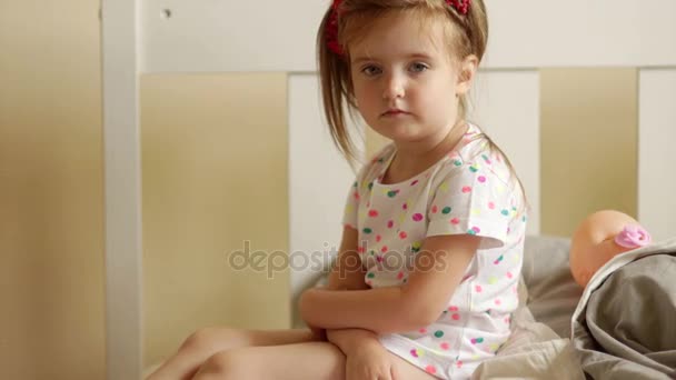 Serious child looking at camera sits on bed. Behind her is her doll. Favorite toy. Close-up portrait — Stock Video