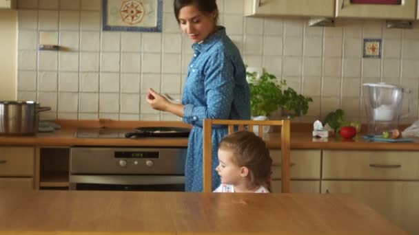 Maman cuisine des œufs sur la cuisinière. Une petite fille attend le petit déjeuner à table. La fille est très en colère. Des caprices enfantins. Petit déjeuner familial . — Video