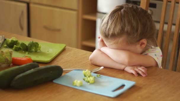 Retrato de uma menina na cozinha. A mãe dá à filha um brócolis e uma cenoura. A rapariga afasta os vegetais. Comida saudável. Caprichos infantis . — Vídeo de Stock