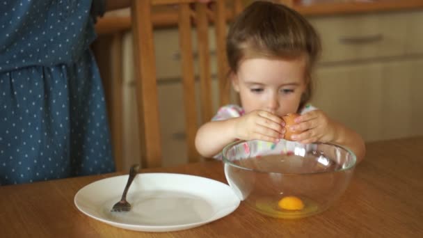 Gesloten portret van een meisje in de keuken. De dochter zelf breekt de eieren in een kom. Moeder veegt haar handen met een handdoek. Moeders helper. — Stockvideo