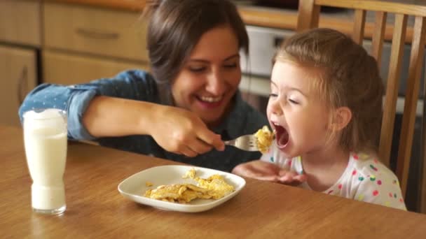 Belle brune nourrit sa fille. La fille ouvre grand la bouche. Maman rit. Œufs brouillés et lait. Petit déjeuner . — Video