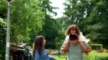 Anne, baba ve kızı cadde boyunca yürüyor. Kızı baba omuz oturur. Neşeyle gülüyorlar.