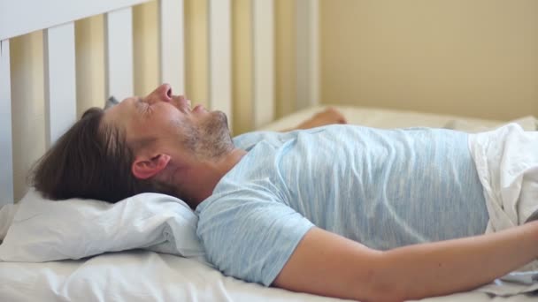 Unrasierter Mann, der aus dem Schlaf auf einem bequemen Bett aufwacht. er nimmt Deckung mit einer Decke und wendet sich auf der anderen Seite ab, um zu schlafen — Stockvideo