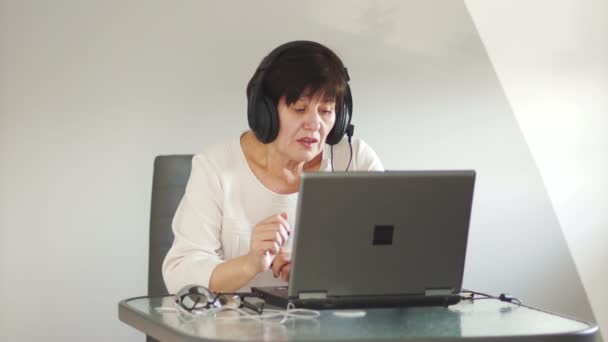 带麦克风的妇女通过电脑与客户交谈。妇女与个人计算机通过 skype 通信。有手提电脑的女人资深妇女与耳机和笔记本讲话与顾客 — 图库视频影像