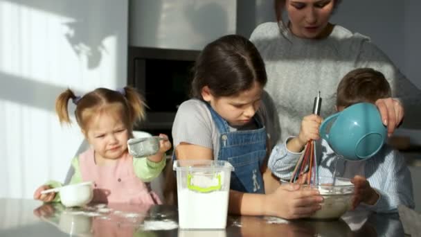 Трое детей разного возраста смешивают тесто. Старшая сестра муки в миске. Мама на заднем плане управляет процессом . — стоковое видео