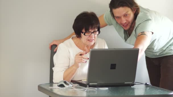 Un joven ayuda a su madre a aprender una computadora. Pacientemente le muestra y le explica el procedimiento de las acciones — Vídeo de stock