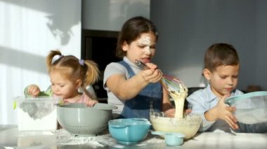 Üç çocuk mutfakta. Kız hamur kıpırdanırken ve izliyor akışı corolla. Anne onları un elemek yardımcı olur