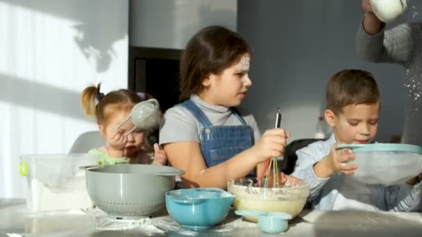 Desorden en la cocina. Tres niños alegres amasan la masa para pasteles. Mamá les ayuda a tamizar la harina. Cocina familiar. Corola para masa, tamiz, tazón, utensilios de cocina — Vídeo de stock