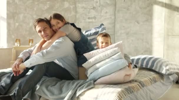 Un padre joven con dos hijos está sentado en la cama. Mañana de una familia numerosa. Chica abrazando padre, chico jugando con almohadas — Vídeos de Stock