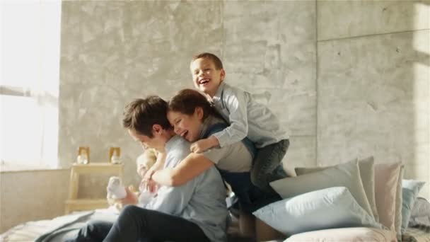 Ein junger Vater und drei Kinder umarmen sich auf dem Bett sitzend. Glückliche Vaterschaft. Vatertag — Stockvideo