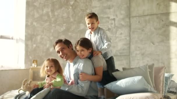 Tres hijos y un padre joven abrazan sentarse en la cama. Mañana familiar. Feliz paternidad. Día del Padre — Vídeos de Stock