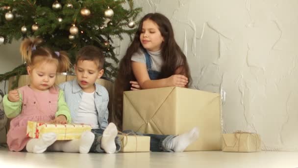 Tre barn öppnar julklapp tillsammans. Barnet själv tar paketet av gåvan — Stockvideo